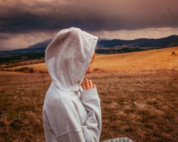 Person meditating in hoodie outdoors with mountains and cloudy sky in view.